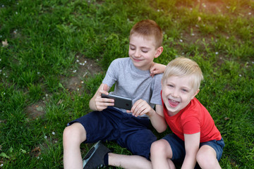Naklejka na ściany i meble Two boys with a smartphone in their hands are sitting on the grass.
