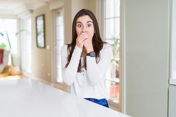 Beautiful young woman sitting on white table at home shocked covering mouth with hands for mistake. Secret concept.