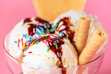 scoops of ice cream in glass bowl with chocolate sauce, strewed sprinkles and cookies on pink background, close up