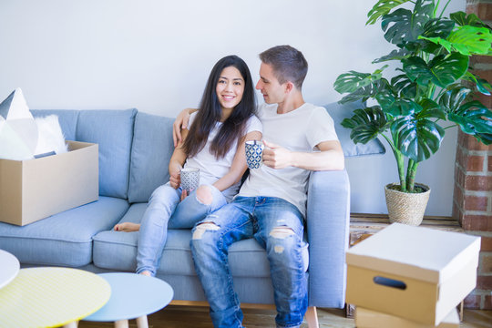 Young beautiful couple sitting on the sofa drinking coffee at new home around cardboard boxes