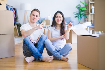 Fototapeta na wymiar Young beautiful couple sitting on the floor at new home around cardboard boxes with a big smile on face, pointing with hand and finger to the side looking at the camera.