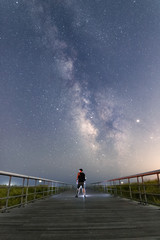 Silhouette of a man standing on a long boardwalk leading to the ocean with the Milky Way galactic core shining brightly in the sky. 