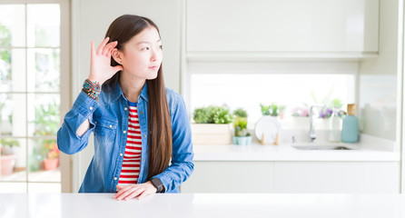 Beautiful Asian woman wearing denim jacket on white table smiling with hand over ear listening an hearing to rumor or gossip. Deafness concept.