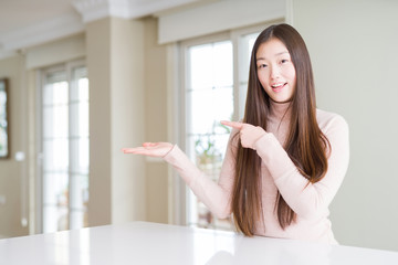 Beautiful Asian woman wearing casual sweater on white table amazed and smiling to the camera while presenting with hand and pointing with finger.