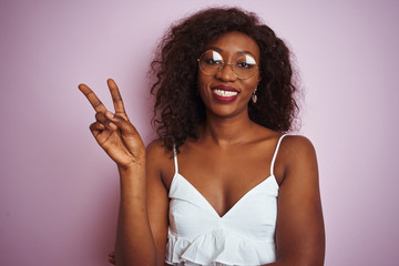 Young african american woman wearing glasses standing over isolated pink background smiling with happy face winking at the camera doing victory sign. Number two.