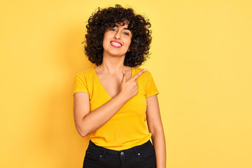 Young arab woman with curly hair wearing t-shirt standing over isolated yellow background cheerful with a smile on face pointing with hand and finger up to the side with happy and natural expression