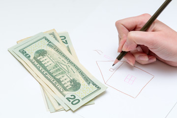 woman draws a pencil house on a sheet of paper. Next to it is a dollar bill. The concept is a mortgage. Dream of own housing