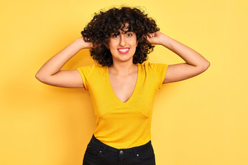 Young arab woman with curly hair wearing t-shirt standing over isolated yellow background Smiling pulling ears with fingers, funny gesture. Audition problem