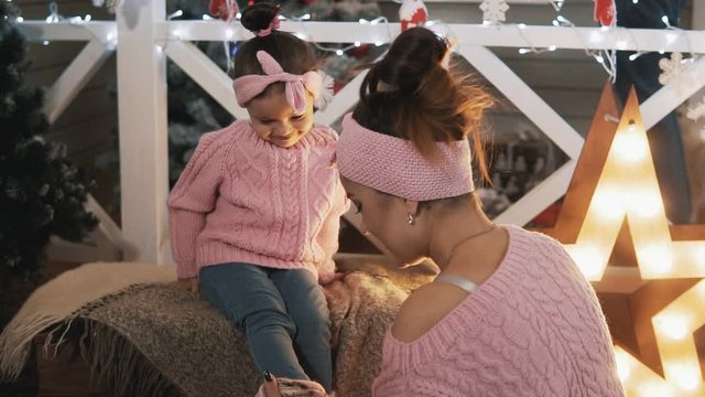 Beautiful mother and daughter in pink pullovers. Happy mother having fun with her kid daughter near Christmas tree. Mother tickling her little daughter in slow motion. Motherhood concept. Medium shot