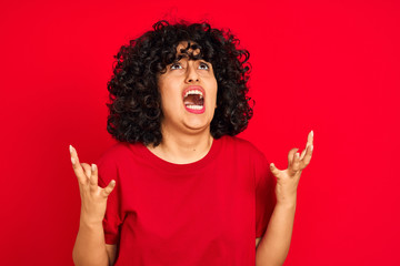 Young arab woman with curly hair wearing casual t-shirt over isolated red background crazy and mad shouting and yelling with aggressive expression and arms raised. Frustration concept.