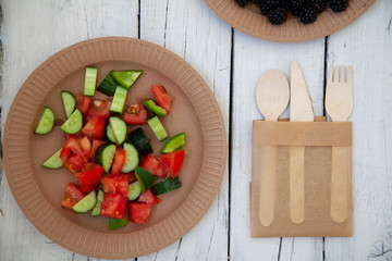 paper cardboard dishes with vegetable salad tomatoes cucumbers with wooden devices fork horse knife on a white wooden table