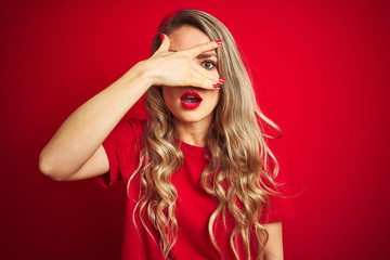 Fototapeta premium Young beautiful woman wearing basic t-shirt standing over red isolated background peeking in shock covering face and eyes with hand, looking through fingers with embarrassed expression.