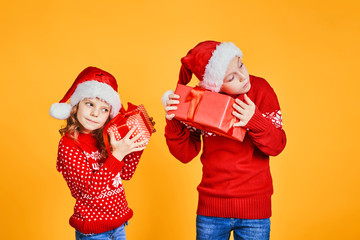 Cheerful kids in Santa hats and red sweaters standing with presents checking what is inside of gift box on yellow background