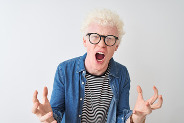 Young albino blond man wearing denim shirt and glasses over isolated white background crazy and mad shouting and yelling with aggressive expression and arms raised. Frustration concept.