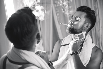 Man looking in the mirror, smoking a cigarette and shaving his beard with a straight razor