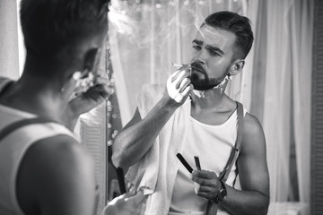 Man looking in the mirror, smoking a cigarette and shaving his beard with a straight razor