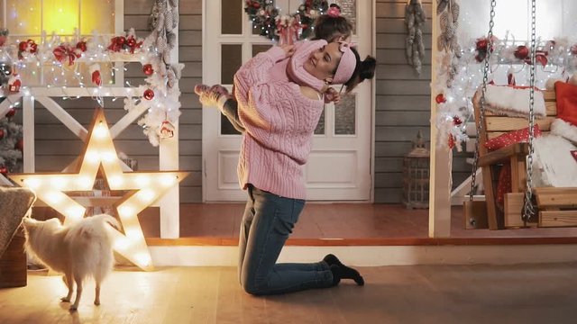 Beautiful mother and daughter in pink pullovers at Christmas day. Happy mother having fun with her kid daughter at Christmas porch. Mother tickling her little daughter in slow motion. Full length shot