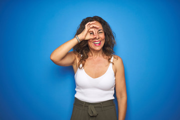 Fototapeta premium Middle age senior woman with curly hair standing over blue isolated background doing ok gesture with hand smiling, eye looking through fingers with happy face.