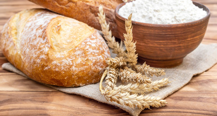 Loafs of bread with wheat ears and bowl with flour on the wooden table.