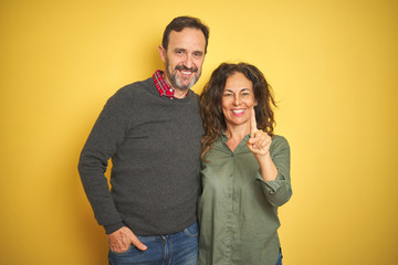 Fototapeta premium Beautiful middle age couple over isolated yellow background showing and pointing up with finger number one while smiling confident and happy.