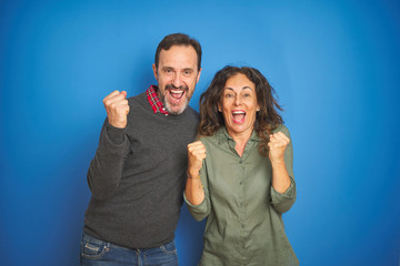 Beautiful middle age couple together standing over isolated blue background celebrating surprised...