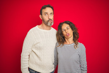 Fototapeta premium Beautiful middle age couple wearing winter sweater over isolated red background making fish face with lips, crazy and comical gesture. Funny expression.