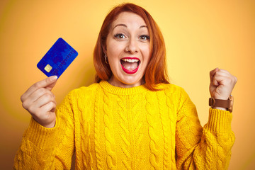 Young customer redhead woman holding credit card over yellow isolated background screaming proud and celebrating victory and success very excited, cheering emotion