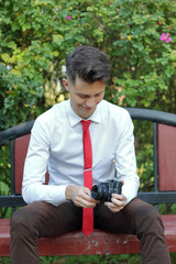 A stylishly dressed young man sits on a park bench. Stylishly dressed young man sits on a park bench. In his hands holds a SLR film camera.