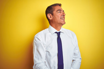 Handsome middle age businessman standing over isolated yellow background looking away to side with smile on face, natural expression. Laughing confident.
