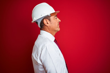 Handsome middle age engineer man wearing security helmet over isolated red background looking to side, relax profile pose with natural face with confident smile.