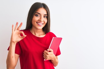 Young beautiful student woman holding red book standing over isolated white background doing ok sign with fingers, excellent symbol