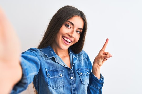 Beautiful woman wearing denim shirt make selfie by camera over isolated white background with a big smile on face, pointing with hand finger to the side looking at the camera.