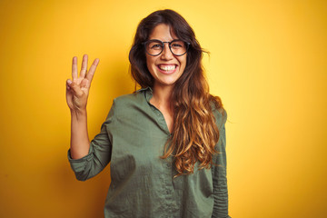 Young beautiful woman wearing green shirt and glasses over yelllow isolated background showing and pointing up with fingers number three while smiling confident and happy.