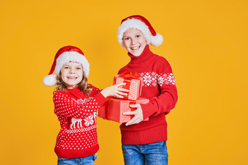 Cheerful kids in Santa hats and red sweaters standing with presents in gift boxes on yellow background