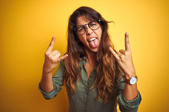 Young beautiful woman wearing green shirt and glasses over yelllow isolated background shouting with crazy expression doing rock symbol with hands up. Music star. Heavy concept.