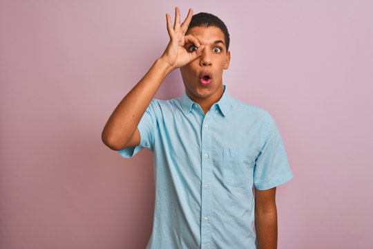 Young handsome arab man wearing blue shirt standing over isolated pink background doing ok gesture shocked with surprised face, eye looking through fingers. Unbelieving expression.