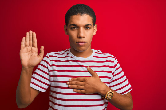 Young handsome arab man wearing striped t-shirt over isolated red background Swearing with hand on chest and open palm, making a loyalty promise oath