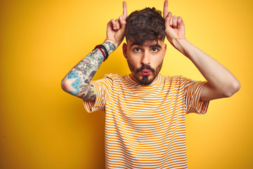 Young man with tattoo wearing striped t-shirt standing over isolated yellow background doing funny...
