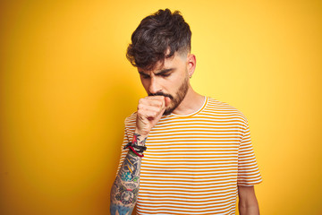 Young man with tattoo wearing striped t-shirt standing over isolated yellow background feeling unwell and coughing as symptom for cold or bronchitis. Healthcare concept.