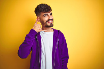 Young man with tattoo wearing sport purple sweatshirt over isolated yellow background smiling doing phone gesture with hand and fingers like talking on the telephone. Communicating concepts.