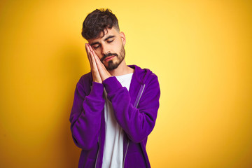 Fototapeta premium Young man with tattoo wearing sport purple sweatshirt over isolated yellow background sleeping tired dreaming and posing with hands together while smiling with closed eyes.