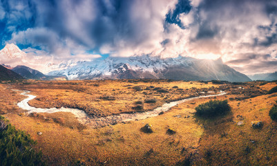 Panoramic landscape with mountains with snow covered peaks in purple clouds, sun, small river, orange grass, blue sky at sunrise. Colorful scenery with meadows, creek, snowy rocks. Autumn in Nepal