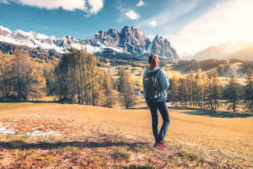 Beautiful young woman with backpack is standing on the hill against mountains at sunset in autumn. Landscape with sporty girl, rocks with snowy peaks, meadow,  orange trees, blue sky in Italy. Travel