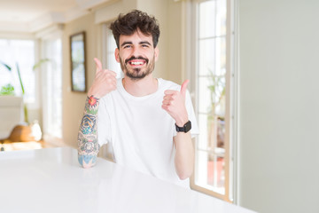 Young man wearing casual t-shirt sitting on white table success sign doing positive gesture with hand, thumbs up smiling and happy. Looking at the camera with cheerful expression, winner gesture.