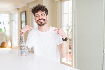 Young man wearing casual t-shirt sitting on white table looking confident with smile on face, pointing oneself with fingers proud and happy.