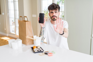 Young man eating asian sushi from home delivery and showing smartphone screen with open hand doing stop sign with serious and confident expression, defense gesture