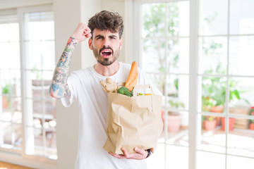 Young man holding paper bag of fresh groceries from the supermarket annoyed and frustrated shouting with anger, crazy and yelling with raised hand, anger concept