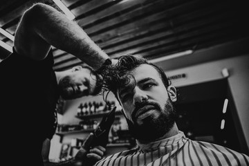 A stylish bearded client sits in a chair, a muscular barber cuts his black hair and makes a hairdo. Selective focus, noise effect