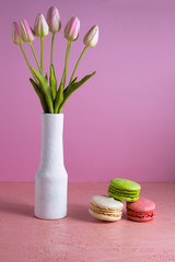 Macaroons on a pale pink background next to a vase of tulips. Place for text.