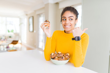 Young african american woman eating chocolate chips muffins pointing and showing with thumb up to the side with happy face smiling